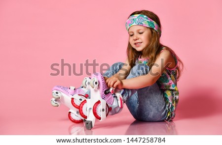 Little baby blond hair child sitting with roller skates in a white shirt and hairband happy smile on a pink studio background.