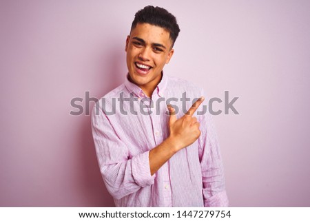 Young brazilian man wearing shirt standing over isolated pink background cheerful with a smile of face pointing with hand and finger up to the side with happy and natural expression on face