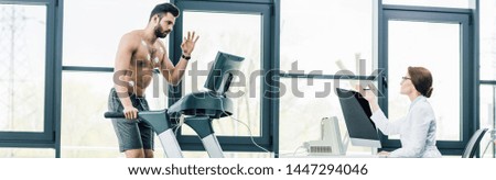 panoramic shot of sportsman gesturing on treadmill near doctor during endurance test in gym