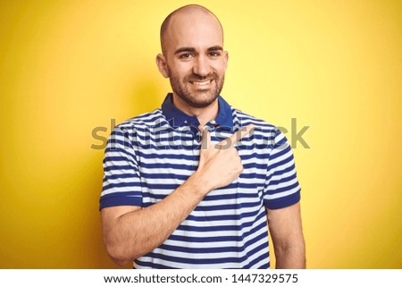 Young bald man with beard wearing casual striped blue t-shirt over yellow isolated background cheerful with a smile of face pointing with hand and finger up to the side 