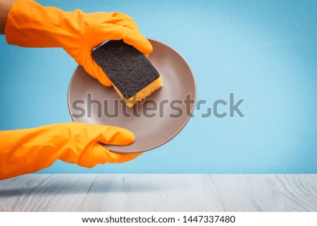 Women's hands in orange protective glove with plate and sponge on gray and blue background. Washing and cleaning concept.