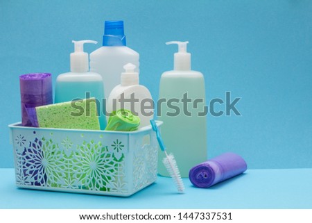 Plastic bottles of dishwashing liquid, glass and tile cleaner in basket, sponges, garbage bags and brushes on blue background. Washing and cleaning concept.