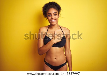 African american woman on vacation wearing bikini standing over isolated yellow background cheerful with a smile on face pointing with hand and finger up to the side with happy and natural expression