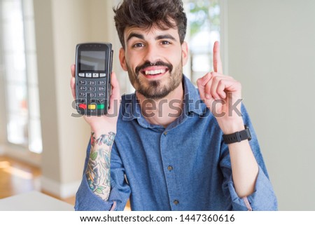 Young man holding dataphone point of sale as payment surprised with an idea or question pointing finger with happy face, number one