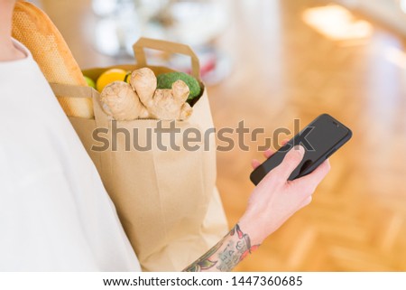 Young handsome man holding a paper bag full of fresh groceries at home, showing and using smartphone while buying products