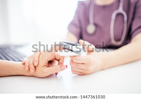 Doctor in purple uniform and shethoskope on a neck holds patient's hand with oximeter at patient's finger. White table. Medical concept. Royalty-Free Stock Photo #1447361030