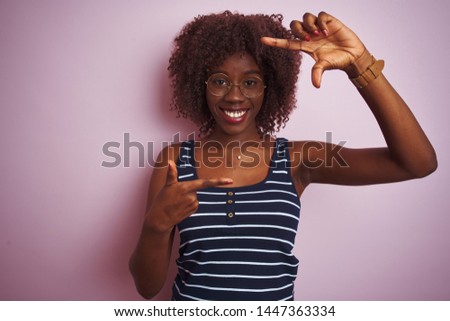 Young african afro woman wearing striped t-shirt glasses over isolated pink background smiling making frame with hands and fingers with happy face. Creativity and photography concept.