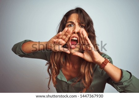 Young beautiful woman wearing green shirt standing over grey isolated background Shouting angry out loud with hands over mouth