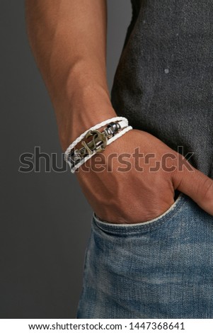 Cropped from top and bottom view shot of a man with a bracelet on his arm. The fashion model on the gray background is dressed in a gray shirt and jeans. You can see a multi-layer bracelet made of lea