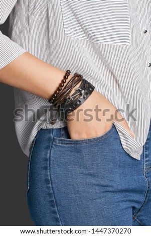 Cropped top and bottom photo of a woman with a bracelet on her arm. The model on a gray background is dressed in a light striped shirt and jeans. You can see a multi-layer bracelet made from leather, 