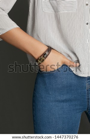 Cropped from top and bottom view shot of a woman with a bracelet on her arm. The fashion model on the gray background is dressed in a light striped shirt and jeans. You can see a multi-layer bracelet 