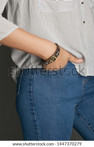 Cropped from top and bottom view shot of a woman with a bracelet on her arm. The fashion model on the gray background is dressed in a light striped shirt and jeans. You can see a multi-layer bracelet 