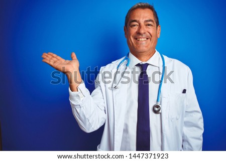 Handsome middle age doctor man wearing stethoscope over isolated blue background smiling cheerful presenting and pointing with palm of hand looking at the camera.