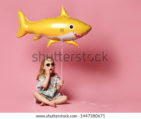little girl in a summer dress, wears a balloon in the shape of a yellow shark-fish, celebrates a holiday, smiles broadly, against a pink background. The concept of children and recreation