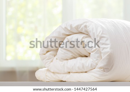 A folded rolls duvet is lying on the dresser against the background of a blurred window. Household. Royalty-Free Stock Photo #1447427564