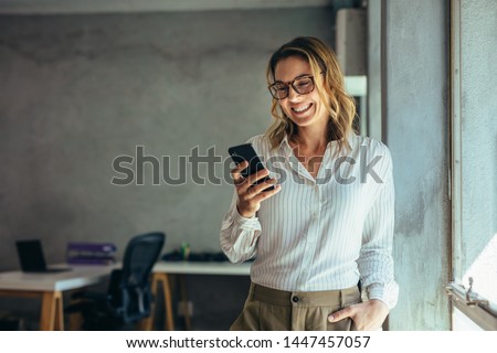 Smiling businesswoman using phone in office. Small business entrepreneur looking at her mobile phone and smiling. Royalty-Free Stock Photo #1447457057