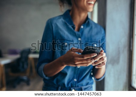 Woman in casuals standing in office by a window with phone in hand. Businesswoman with mobile phone in hand. Royalty-Free Stock Photo #1447457063