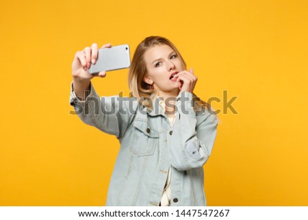 Portrait of pretty young woman in denim casual clothes gnawing nails, doing selfie shot on mobile phone isolated on yellow orange background in studio. People lifestyle concept. Mock up copy space