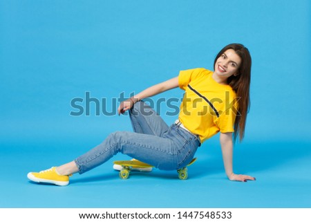 Portrait of charming young woman in vivid casual clothes looking camera and sitting with yellow skateboard isolated on blue wall background in studio. People lifestyle concept. Mock up copy space
