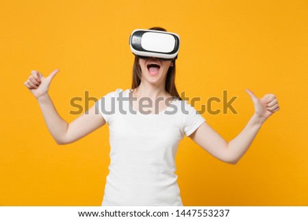 Cheerful young girl in white casual clothes watching in headset of virtual reality pointing thumbs on herself isolated on bright yellow orange background. People lifestyle concept. Mock up copy space
