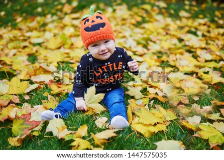 Adorable little girl in Halloween theme clothes sitting on the grass and playing with colorful autumn leaves on a fall day in park