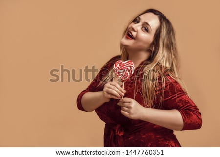 Sending you my love. Plus size happy woman in velour dress holding a red heart-shaped lollipop and looking at camera with smile while standing in studio.