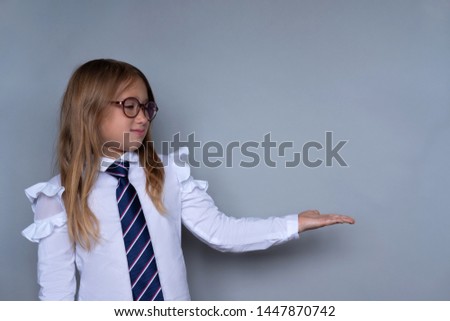 Small schoolchild in glasses showing something with palm of hand. Nerdy schoolgirl presenting school project portrait. Schoolgirl, preteen child with cupped hand in school uniform on grey background