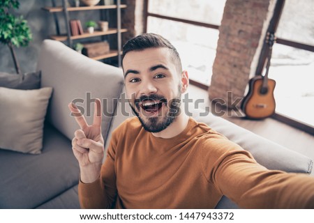 Close up photo of handsome satisfied guy hipster guy make v-signs laughter free time dressed orange pullover sweater sit divan big light room apartment vlog photo