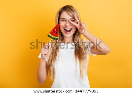 Image of happy pleased positive young blonde woman jumping isolated over yellow wall background dressed in white casual t-shirt holding candy sweeties.