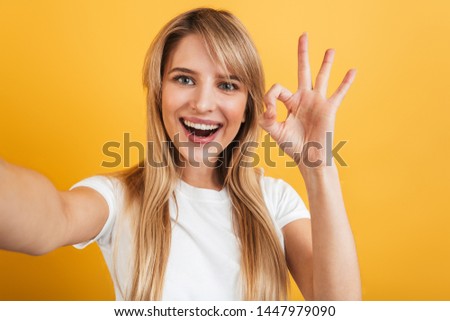 Image of a cheerful smiling positive pleased young blonde woman posing isolated over yellow wall background dressed in white casual t-shirt take selfie by camera showing okay gesture.