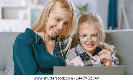 Beautiful Young Mom and Her Cute Little Daugther Use Laptop while Sitting on a Sofa at Home. Family Spending Time Together Watching Videos and Cartoons on Computer.