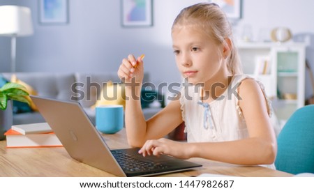 Smart Little Girl Does Homework in Her Living Room. She's Sitting at Her Desk, Uses Laptop and Writes with a Pen in Her Textbooks.
