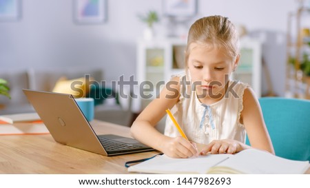 Smart Little Girl Does Homework in Her Living Room. She's Sitting at Her Desk Writes with a Pen in Her Textbooks and  Uses Laptop.