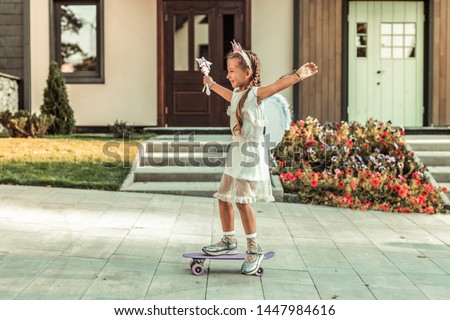 Riding on a skateboard. Long-haired little girl spreading hands while being happy with time spending on an empty street
