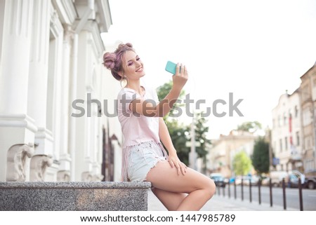 Happy cheerful young blonde with pink hair sitting in the city background taking a picture of herself with a mobile phone. Ginger student girl resting on the street after college