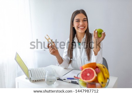 Young female nutritionist sitting at table with fresh vegetables and fruits in her office. Female nutritionist sitting at table with clipboard and healthy products on white background