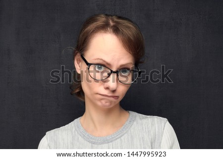 Studio close-up portrait of upset and funny caucasian fair-haired girl with eyeglasses, pouting and biting lips, making naive childish face, looking guiltily at camera, over gray background. Headshot Royalty-Free Stock Photo #1447995923