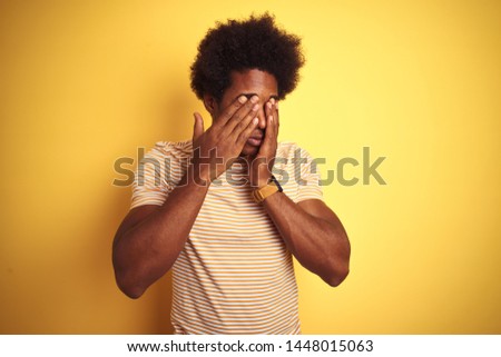 American man with afro hair wearing striped t-shirt standing over isolated yellow background rubbing eyes for fatigue and headache, sleepy and tired expression. Vision problem