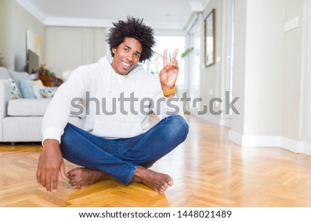 African American man sitting on the floor at home smiling with happy face winking at the camera doing victory sign. Number two.
