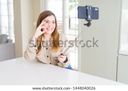 Beautiful young woman taking a picture using selfie stick doing ok sign with fingers, excellent symbol