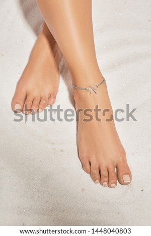 Cropped front shot of girl's legs with french pedicure, wearing silver ankle bracelet, decorated with silver insertion in view of tied bow. The lady is crossing her legs, lying on the sandy platform.