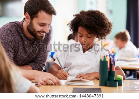 Male Elementary School Teacher Giving Female Pupil Wearing Uniform One To One Support In Classroom Royalty-Free Stock Photo #1448047391