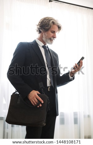 Picture of a concentrated handsome grey-haired senior businessman indoors at home dressed in formal clothes using mobile phone holding bag.