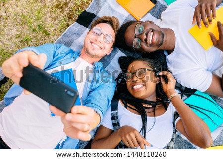Young handsome beard student man taking self-portrait to beautiful black girlfriend with her african brother. Top view. Multiethnic people lying on grass outdoor.