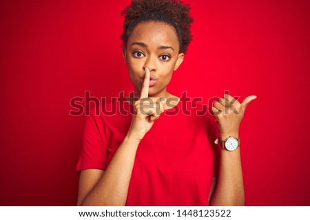 Young beautiful african american woman with afro hair over isolated red background asking to be quiet with finger on lips pointing with hand to the side. Silence and secret concept.