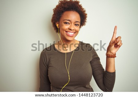 African american woman wearing earphones listening to music over isolated background with a big smile on face, pointing with hand and finger to the side looking at the camera.