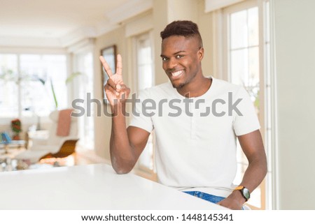 Handsome african american man on white table at home smiling with happy face winking at the camera doing victory sign. Number two.