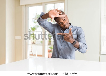 Handsome african american man on white table smiling making frame with hands and fingers with happy face. Creativity and photography concept.