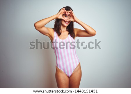 Young beautiful woman wearing striped pink swimsuit swimwear over isolated background Doing heart shape with hand and fingers smiling looking through sign