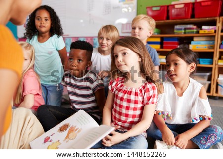 Group Of Elementary School Pupils Sitting On Floor Listening To Female Teacher Read Story Royalty-Free Stock Photo #1448152265